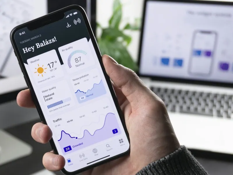 Hand holding a smartphone showing an app data dashboard in front of a design monitor