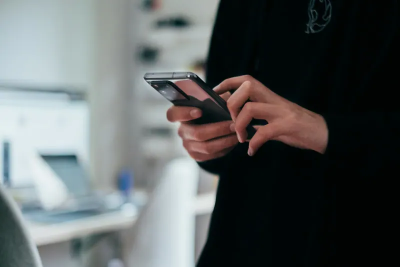 Persona escribe un mensaje en su smartphone en una oficina con computadora al fondo