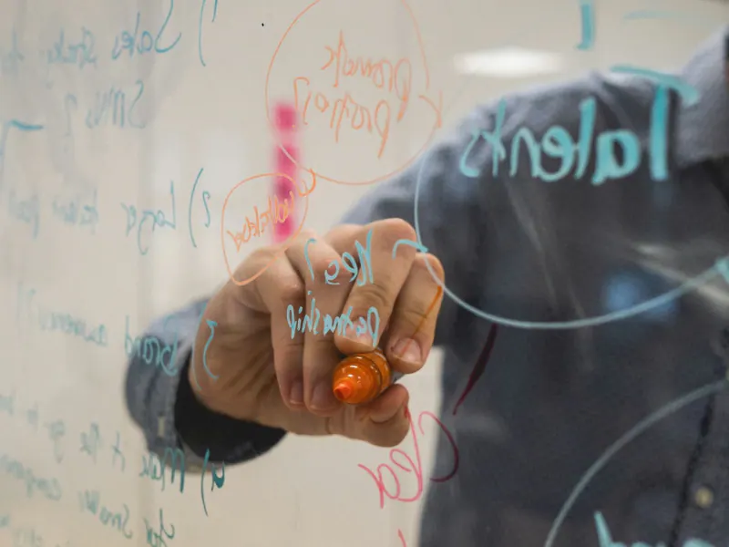 Mano escribe con plumón naranja ideas y diagramas sobre un pizarrón de vidrio durante una sesión de estrategia