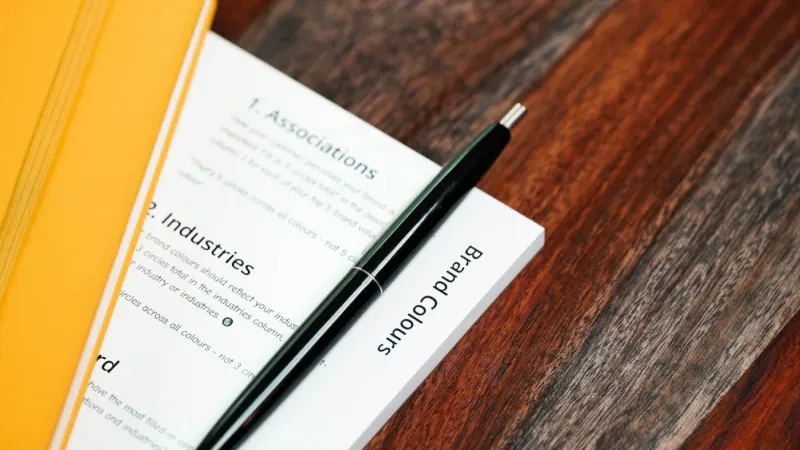 Pluma negra sobre hojas impresas con encabezados, junto a una libreta amarilla
