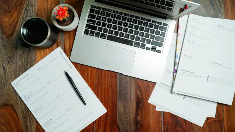 Laptop on a wooden desk with quote documents and a pen