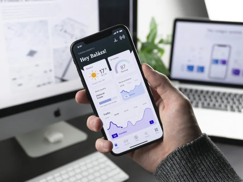 Hand holding a phone with a data dashboard app in front of a computer
