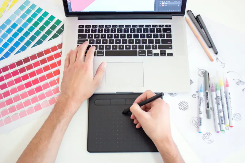 Designer working with a drawing tablet and laptop next to color swatches for a website